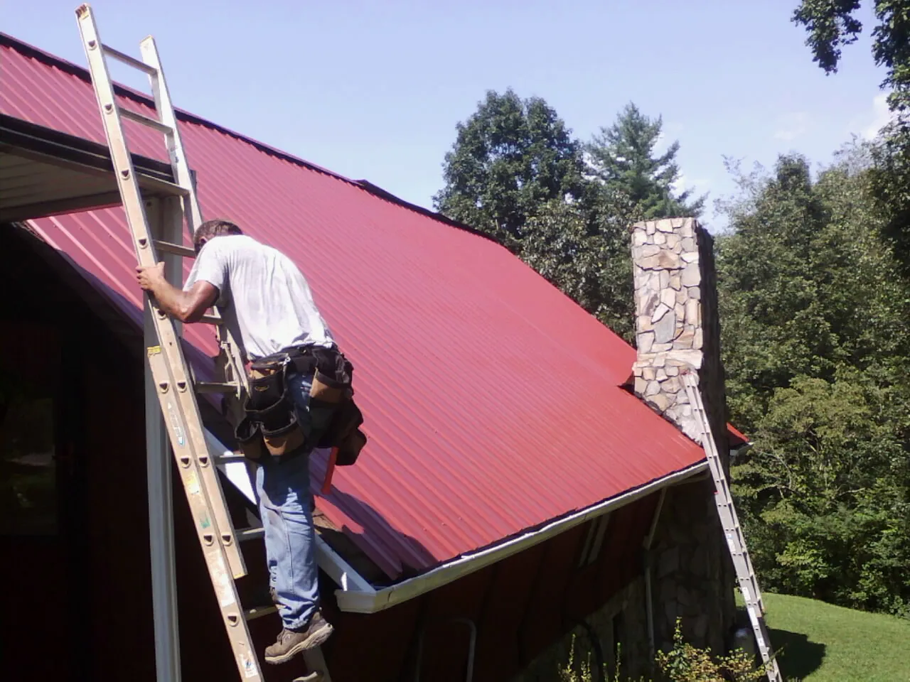 Roofer on a ladder beside a red metal roof and stone chimney — Soffit & Fascia Repair by Roofing Brothers in Clermont